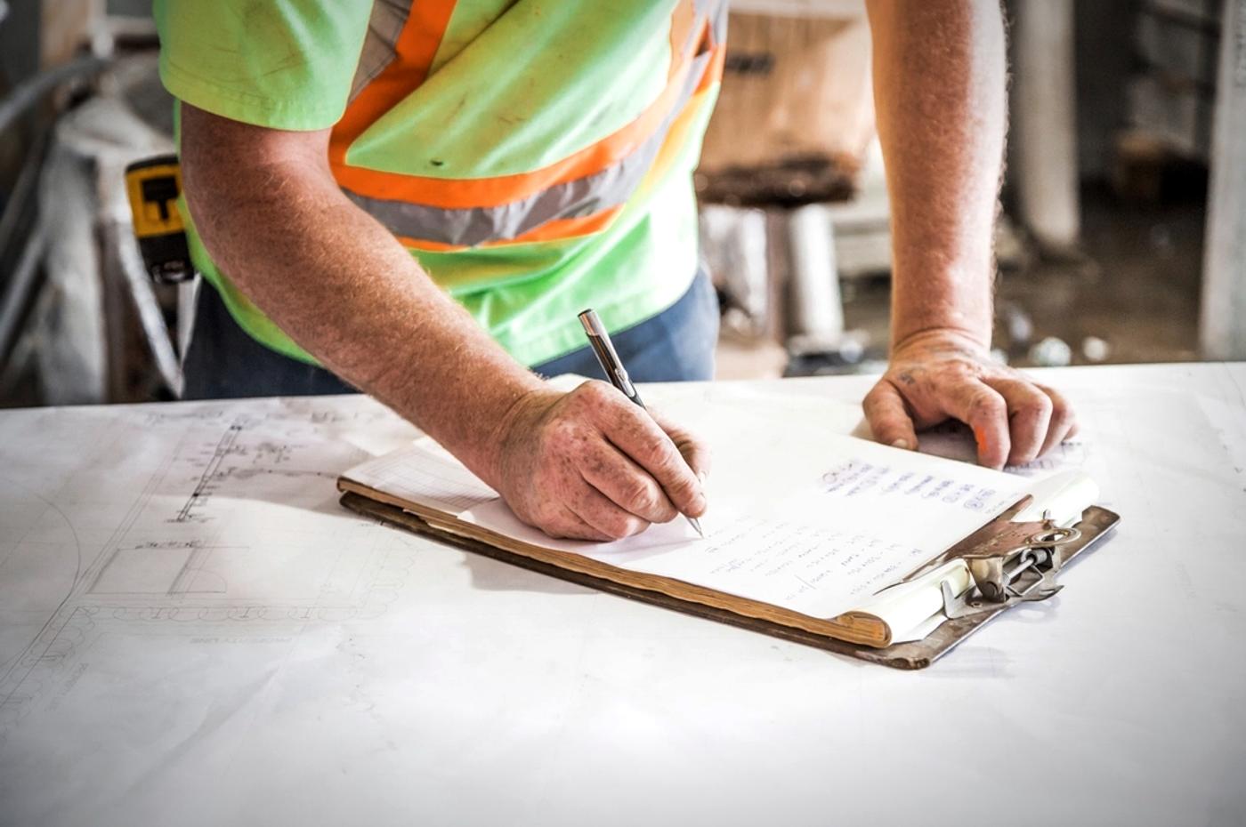Worker in a hi vis vest documenting on a clipboard over blueprints